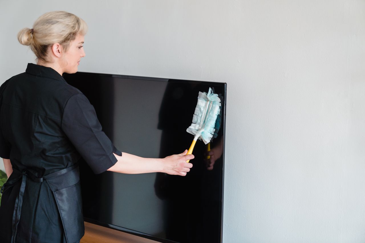 A female housekeeper performing dusting on a television screen indoors.